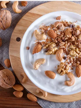 Imagem mostra vasilha de iogurte com granola de castanha-de-caju e coco ao centro sobre tábua de madeiras, e castanhas de caju e amêndoas ao redor da tabua