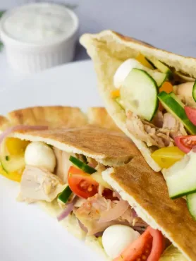 Sanduiche de frango com pao pita aberto mostrando pepino, cebola, tomate e mussarela em bolinhas. Sanduíche em prato branco acompanhado de um potinho com molho